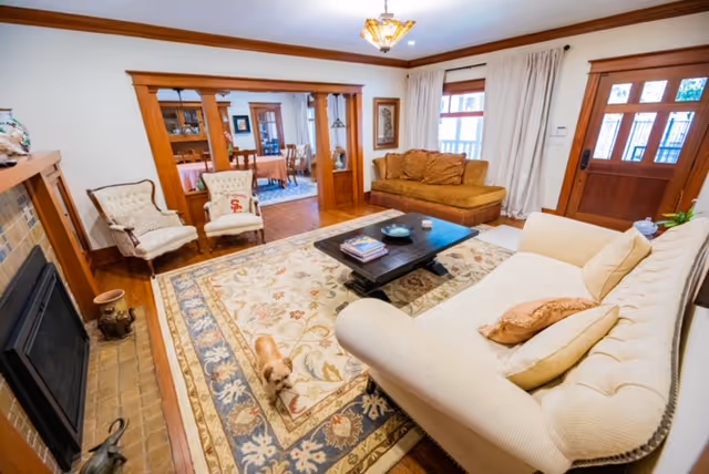 A cozy living room with a cream-colored tufted sofa, a brown cushioned bench, two armchairs, and a dark wooden coffee table on a large patterned area rug. There is a fireplace on the left side and a wooden front door with glass panels on the right. The room has wooden trim and crown molding, with a view into an adjacent dining room through a wide doorway.