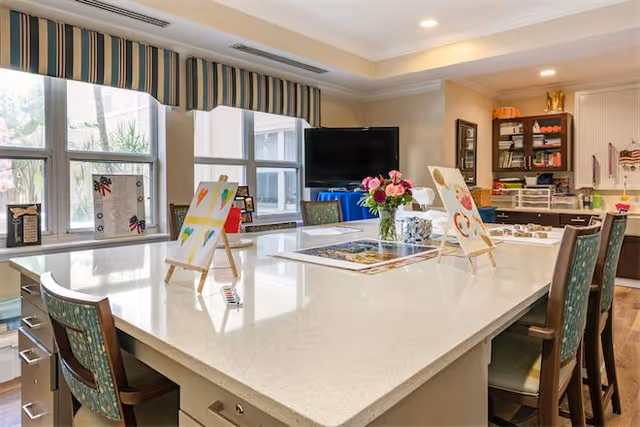 A bright and spacious activity room with a large white countertop island surrounded by chairs. The countertop has small easels displaying colorful paintings, a vase with pink flowers, and various art supplies. Large windows with striped valances allow natural light to fill the room. Shelves and cabinets with organized craft materials are visible in the background.