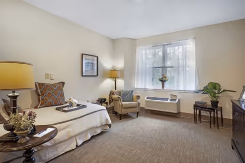 A cozy bedroom in a senior living facility with a single bed covered in a beige blanket and decorative pillows. There is a wooden side table with a lamp and a small plant next to the bed. An armchair with a pillow is placed near a window with sheer white curtains, allowing natural light to fill the room. A small table with a plant and a dresser are also visible.