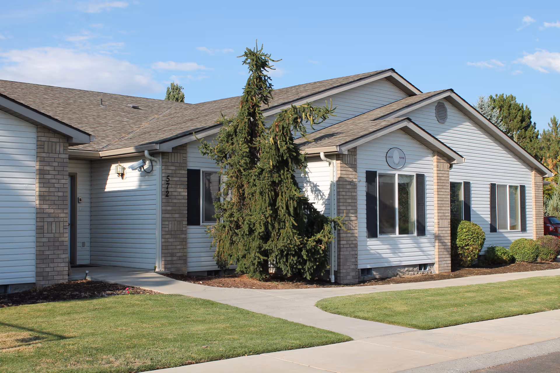 Front exterior of a single-story senior living building with white siding, brick accents, windows, a walkway, and a landscaped lawn.