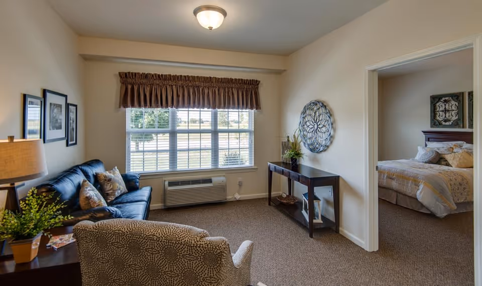 A cozy living room with a patterned armchair and a dark blue sofa adorned with decorative pillows. A wooden side table with a lamp and a plant is next to the sofa. A large window with blinds and a brown valance lets in natural light. A console table with decorative items and a round wall art piece is against the wall. Through an open doorway, a bedroom with a bed, pillows, and wall art is visible.