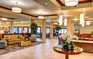 Spacious, well-lit senior living facility common area with seating groups, columns, plants, and a reception desk.