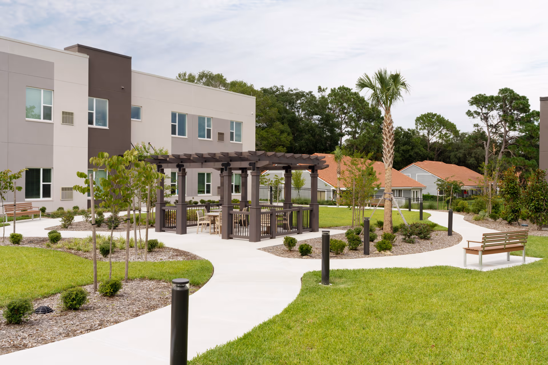 Outdoor garden area at Village on the Green featuring a paved walkway, a wooden pergola with seating underneath, benches, small trees, shrubs, and a palm tree, with residential buildings and greenery in the background.