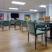 A spacious room with multiple green cushioned chairs arranged around tables and facing a wall-mounted television. The room has light wood flooring, white walls, and a drop ceiling with fluorescent lighting. There are two colorful abstract paintings on the wall and an American flag in the corner. A small sign on one table reads 'Bingo Today'.