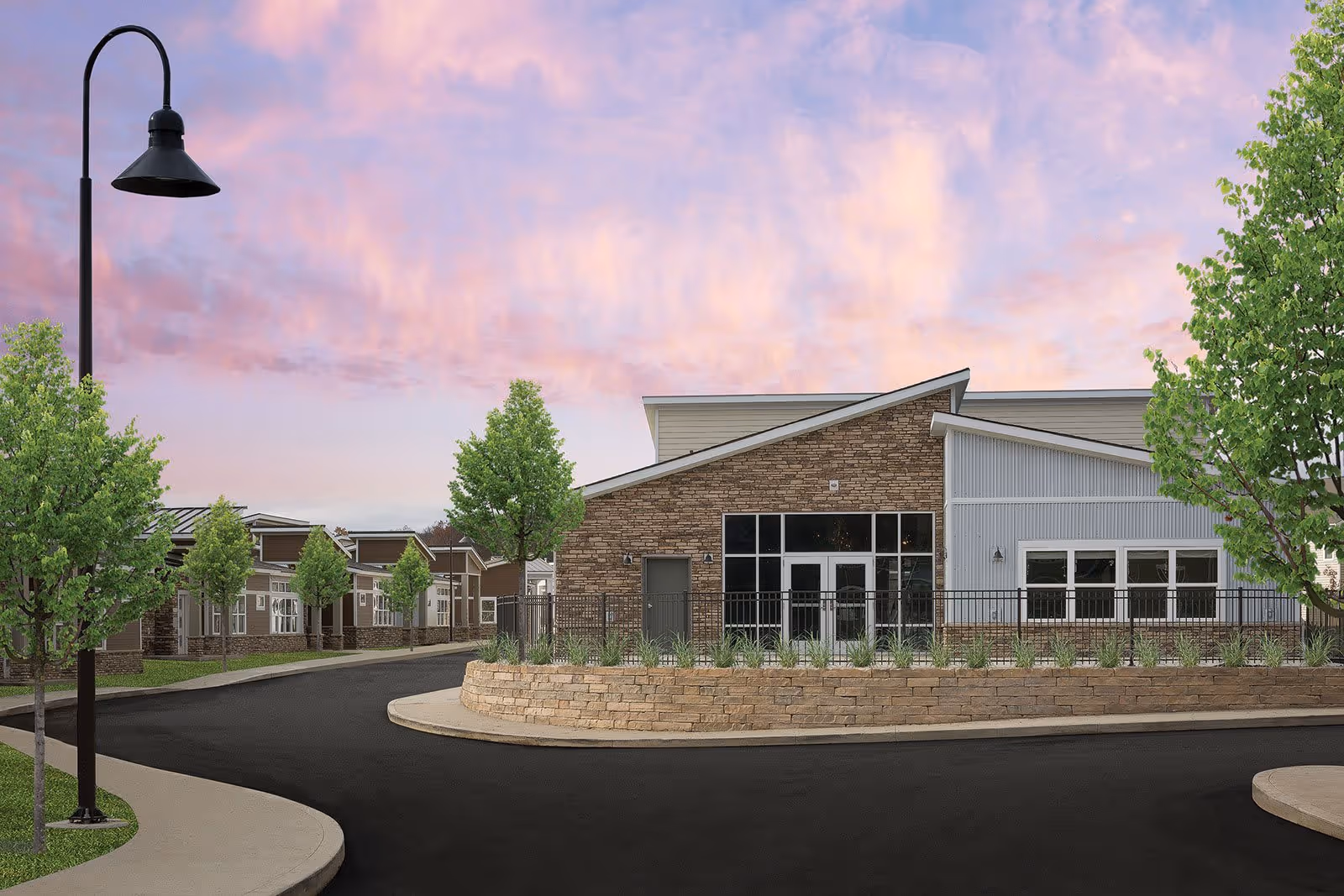 Exterior view of a modern senior living facility building with a combination of brick and light-colored siding, large windows, and a sloped roof. The building is surrounded by a curved paved driveway, landscaped areas with green trees, and a streetlamp. The sky is colorful with a pink and purple sunset.