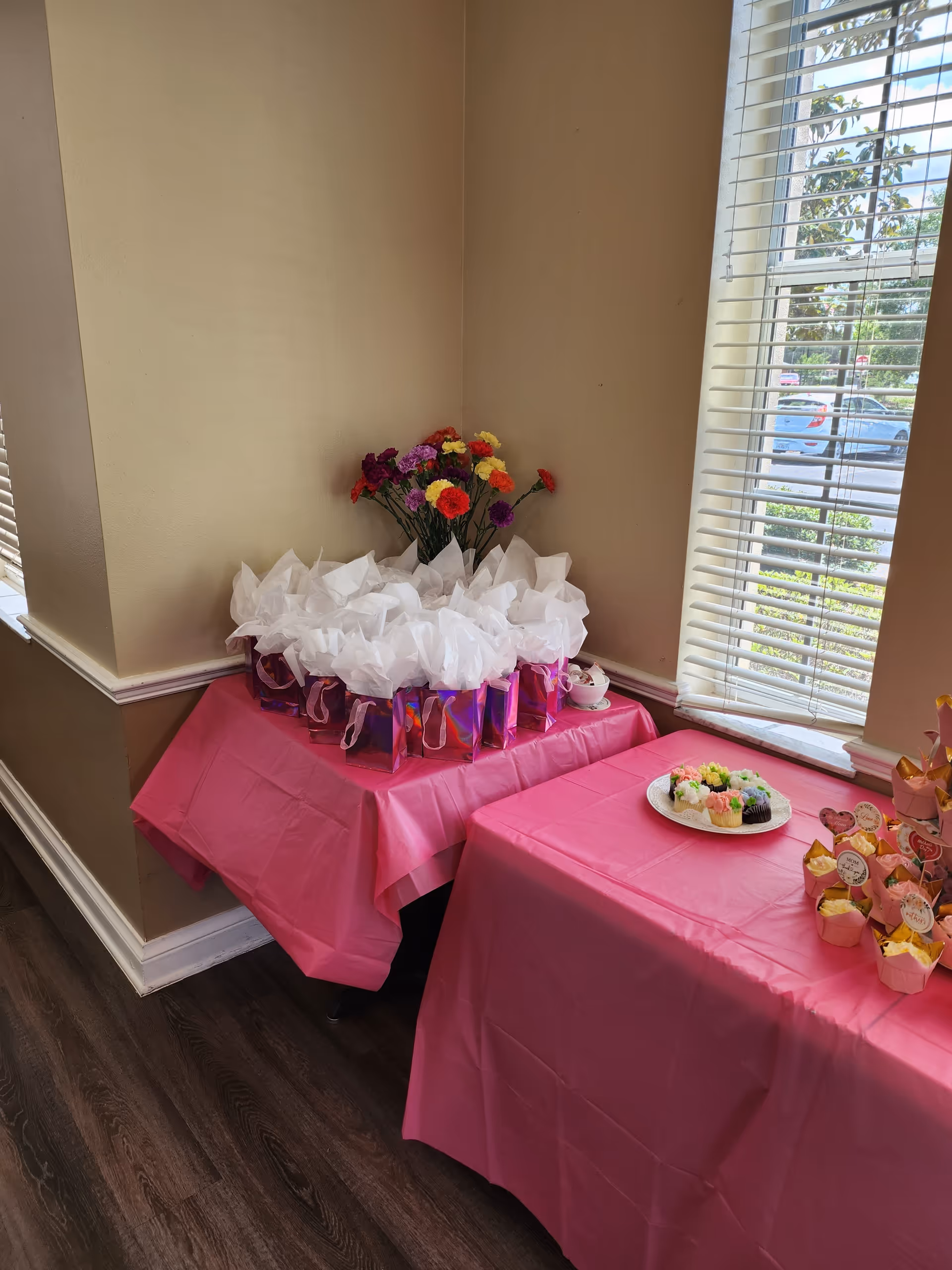 Two tables covered with pink tablecloths set up in a corner near a window with blinds. One table holds several small gift bags with white tissue paper and a vase with colorful flowers. The other table has a plate of decorated cupcakes and more cupcakes with toppers arranged on it. Outside the window, cars and greenery are visible.
