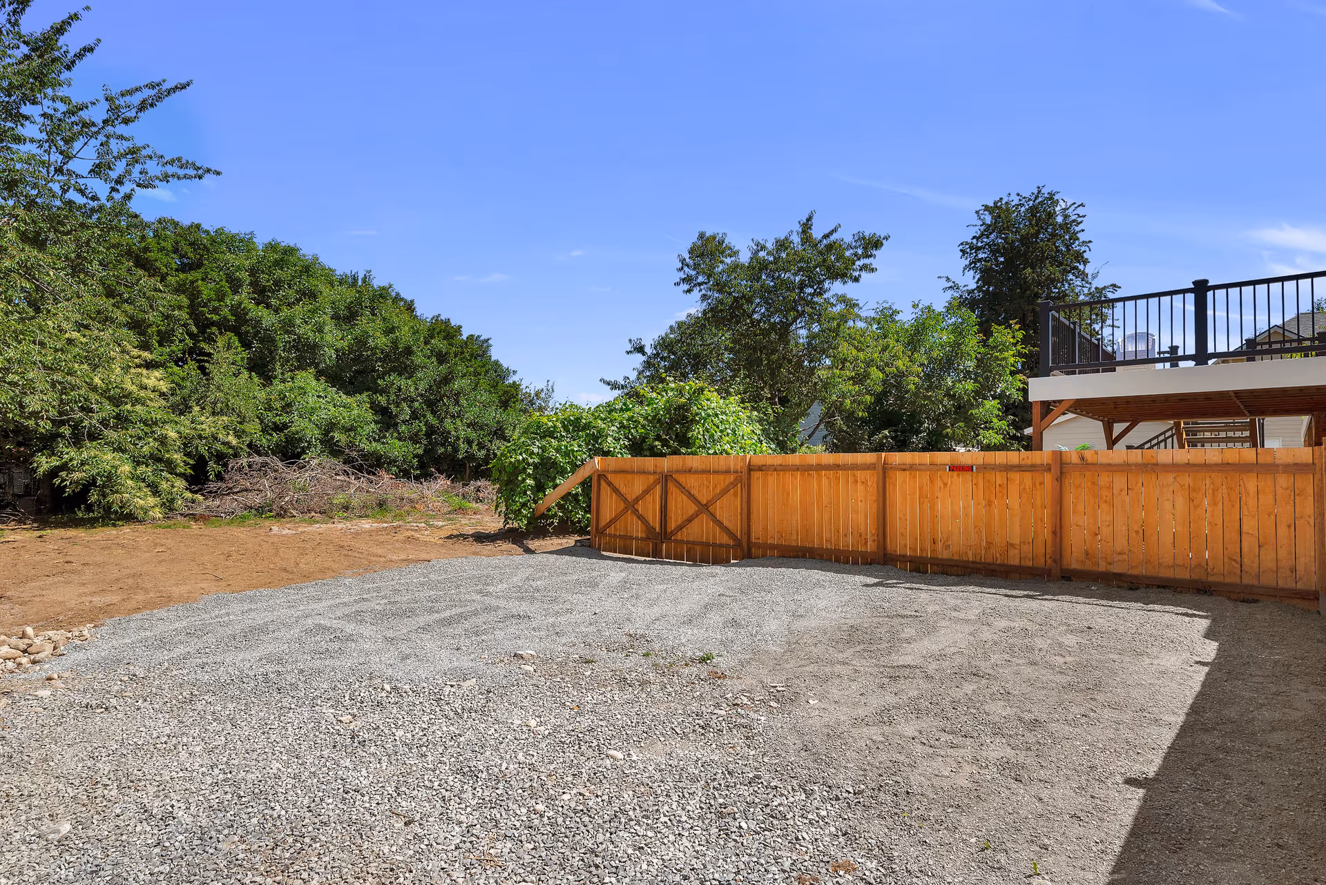 Outdoor area with a gravel ground and a wooden fence. There are green trees and bushes behind the fence, and a clear blue sky above. A raised deck with black railing is visible on the right side.