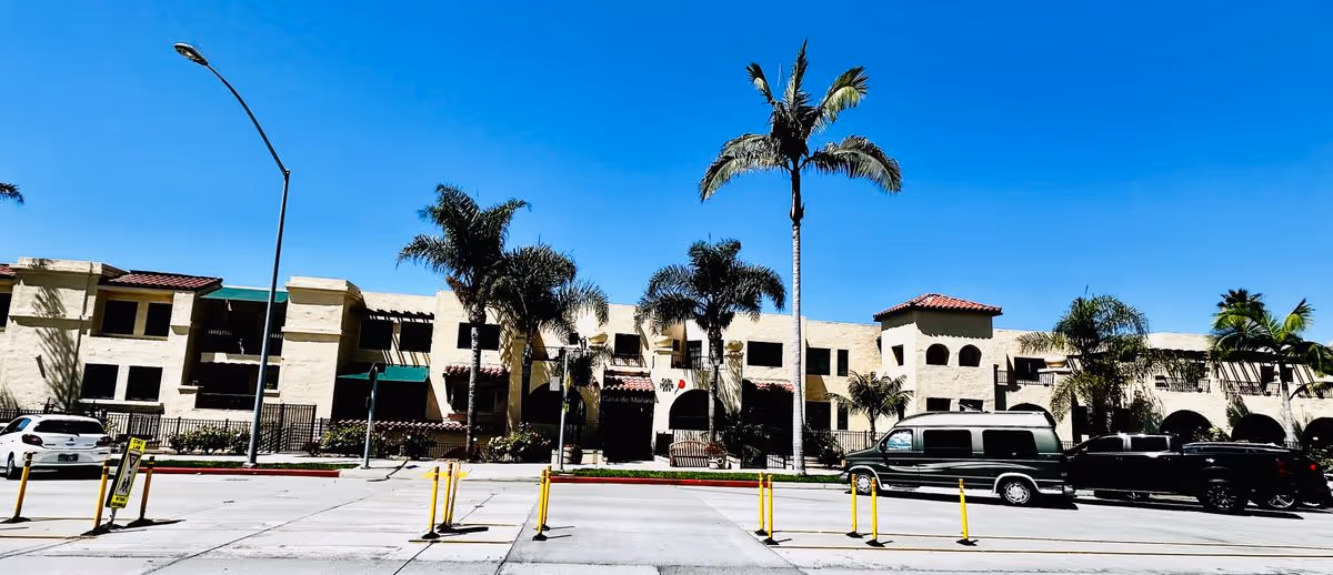 Front view of a two-story Mediterranean-style retirement community building with palm trees, parked vehicles, and a clear blue sky.