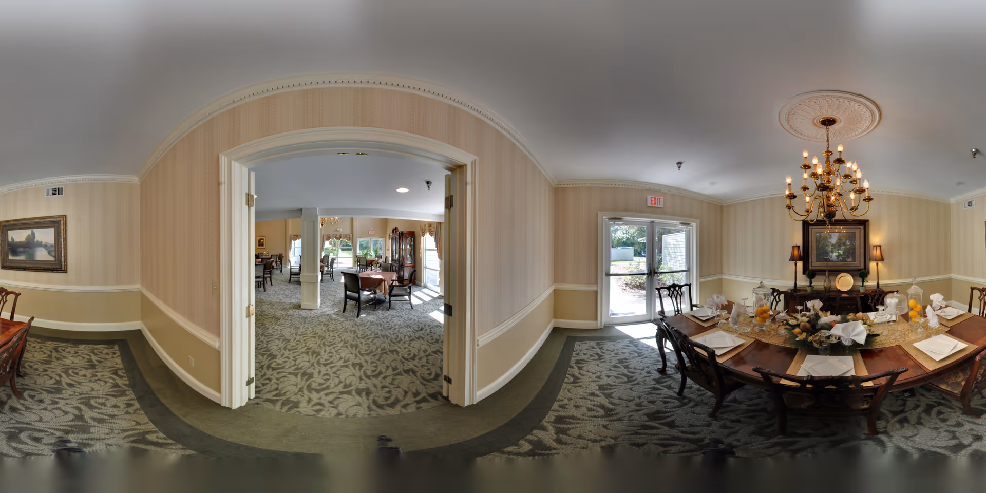 A spacious dining room in a senior living facility with a large wooden dining table set for a meal, including plates, glasses, and napkins. The room features patterned carpet, beige striped wallpaper with white trim, a chandelier hanging from the ceiling, and a sideboard with decorative items and a framed painting. Double doors open to another dining area with additional tables and chairs, and a glass door leads outside, allowing natural light to fill the space.