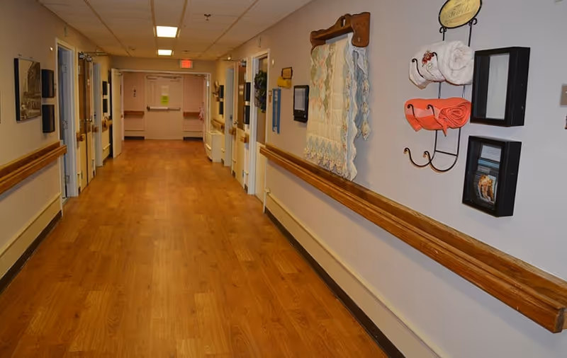 Well-lit interior hallway of a senior care facility with wood floors, handrails, doors, and wall decorations.