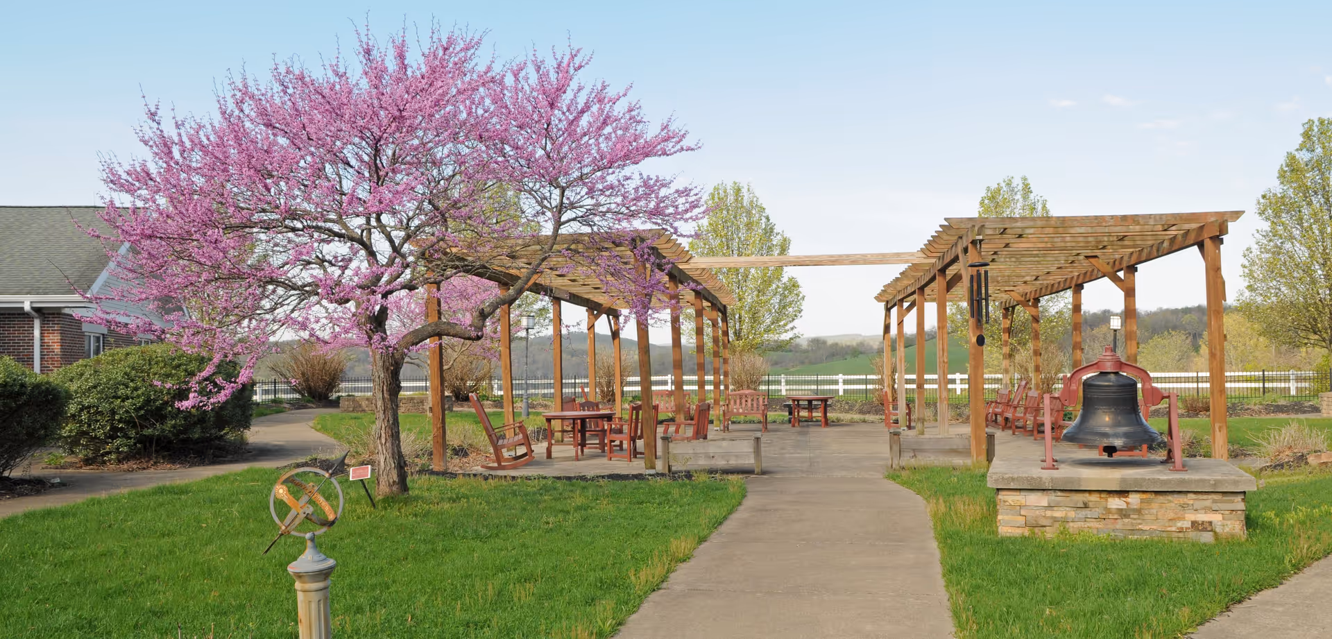Outdoor garden area with a concrete pathway leading between two wooden pergolas. The pergolas have benches and tables underneath. A blooming tree with pink flowers is on the left side, and a large bell mounted on a stone platform is on the right. Green grass and shrubs surround the area with a white fence and trees in the background under a clear sky.