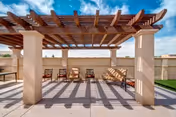 Outdoor seating area with a wooden pergola casting shadows on the concrete floor. The area includes several chairs and a cushioned bench against a beige wall, with a clear blue sky and some clouds visible above.