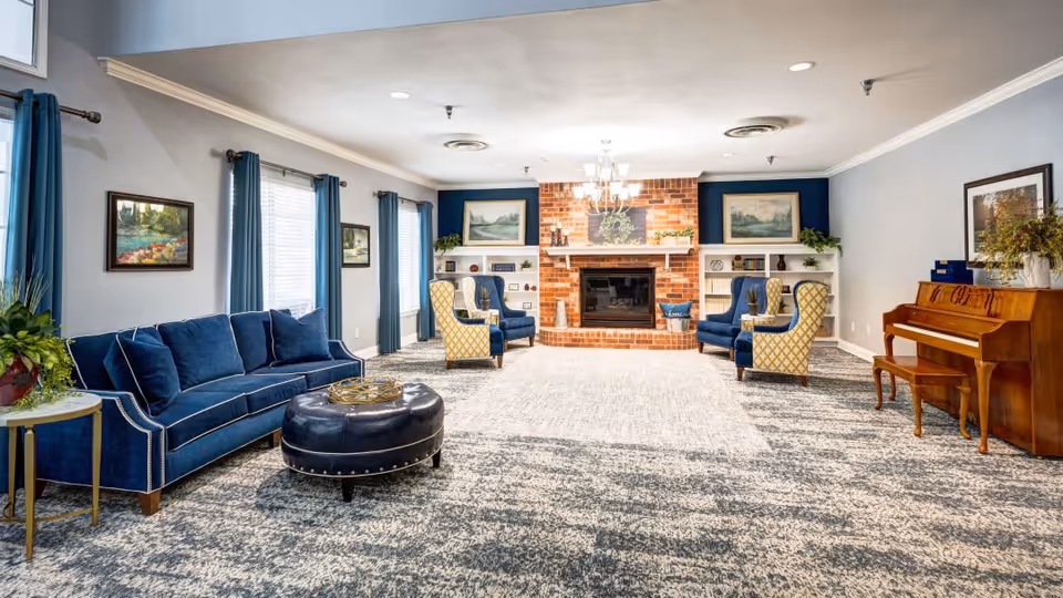 A spacious living room with a patterned carpet, blue velvet sofa, two yellow patterned armchairs, two blue armchairs, a round black leather ottoman, a wooden piano, and a brick fireplace with a chandelier above it. The walls are light gray with blue curtains on the windows and framed paintings hanging on the walls.