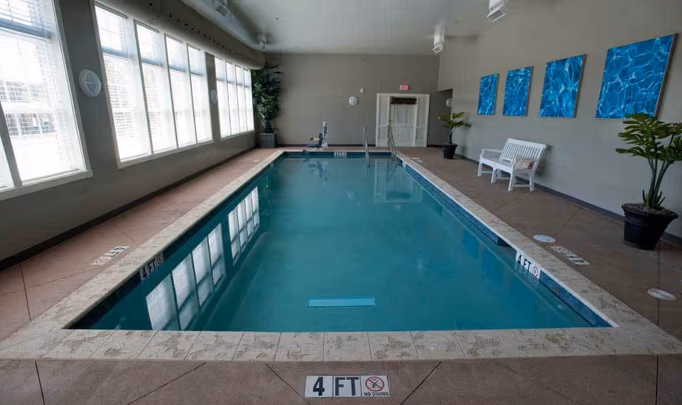 Indoor swimming pool with clear blue water, surrounded by a tiled deck. Large windows line the left side, allowing natural light to fill the space. On the right side, there are three blue water-themed paintings on the wall, a white bench, and potted plants. Depth markers indicate the pool is 4 feet deep with a no diving sign.