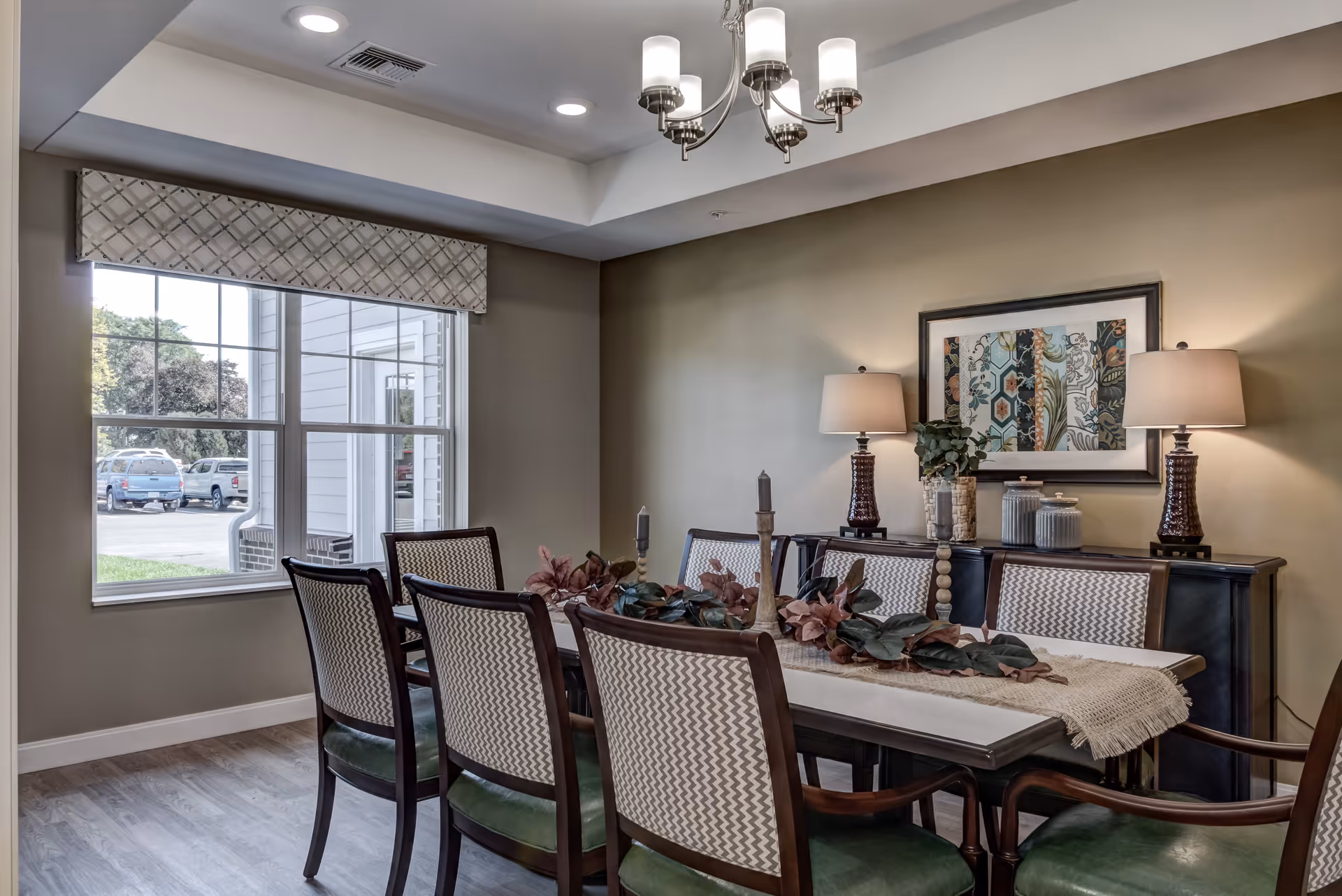 A dining room with a rectangular table surrounded by eight chairs with patterned backs and green seats. The table is decorated with a beige runner and a centerpiece of leaves and candles. Behind the table is a dark sideboard with two matching lamps, decorative jars, and a framed artwork on the wall. A large window with a patterned valance lets in natural light, and the room has a tray ceiling with recessed lighting and a chandelier.