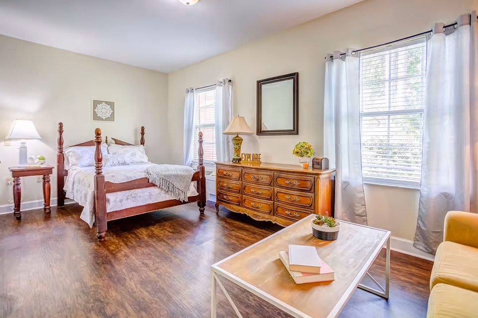 A bright and cozy bedroom with a wooden four-poster bed, white bedding, and a gray throw blanket. There is a wooden nightstand with a lamp and a small plant on the left side of the bed. Two windows with white curtains allow natural light to fill the room. A wooden dresser with a lamp, decorative items, and a mirror above it is against the wall. In the foreground, there is a wooden coffee table with books and a small potted plant, and a yellow armchair is partially visible on the right.