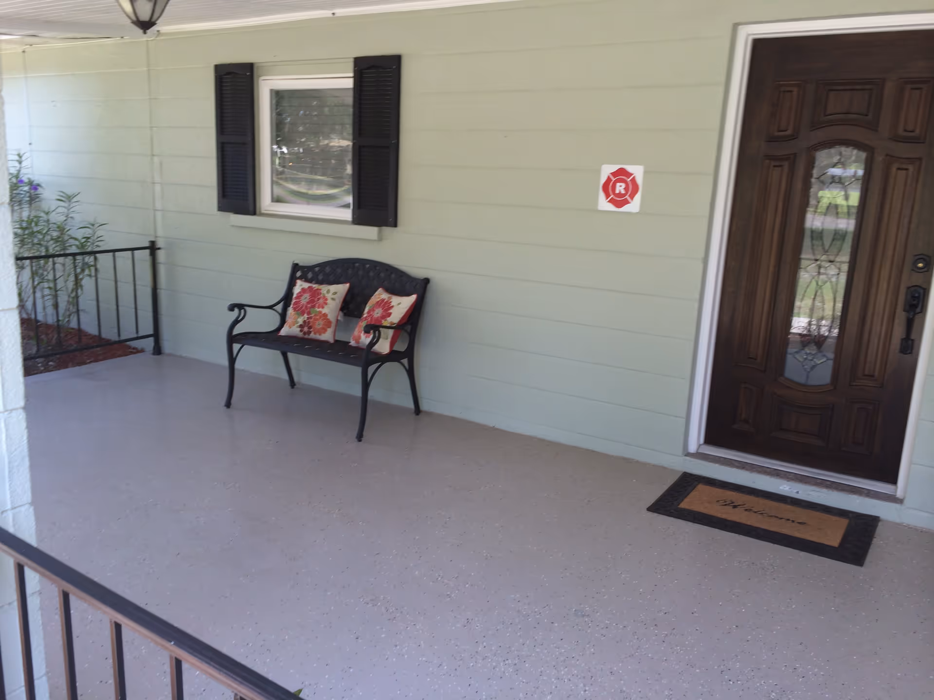 A covered porch area with a light green exterior wall, a dark wooden front door with decorative glass, a black metal bench with two floral cushions, a window with black shutters, a welcome mat, and a small plant in the corner.
