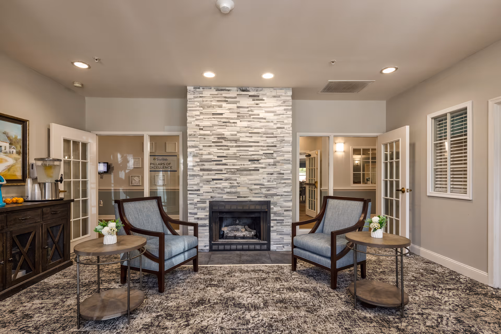 A cozy sitting area with two upholstered armchairs facing a modern stone fireplace. Each chair has a small round wooden side table with a small flower arrangement. The room has a patterned carpet, neutral walls, recessed ceiling lights, and French doors leading to other rooms.