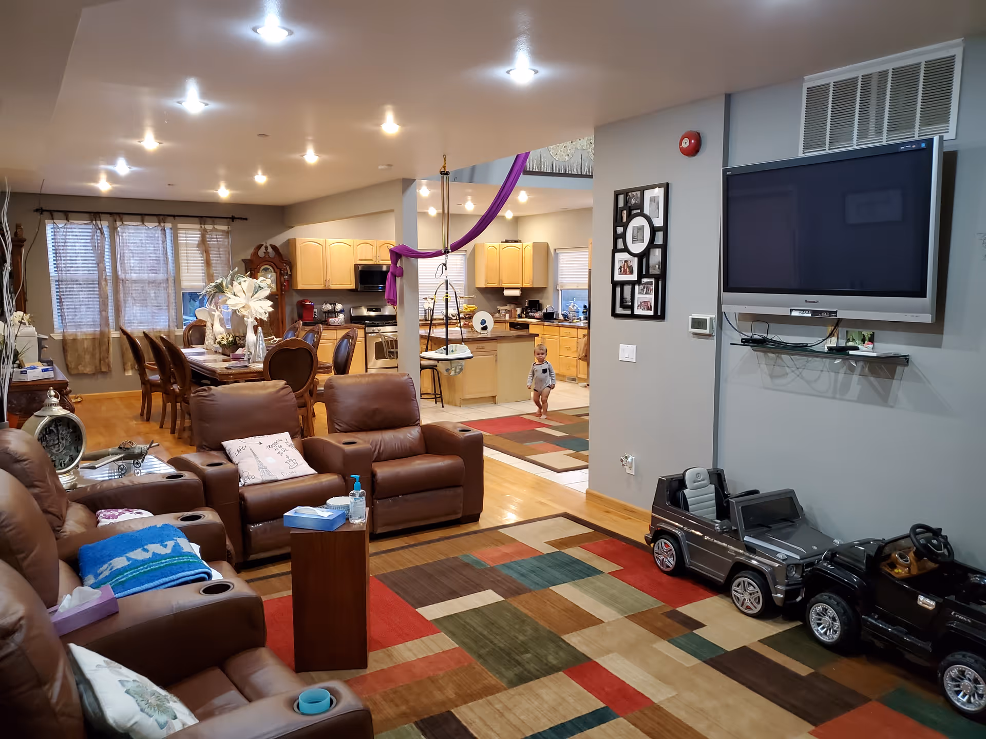 A cozy living room with brown leather recliners, a colorful geometric patterned rug, and a large flat-screen TV mounted on the wall. In the background, there is a dining area with a wooden dining table and chairs, and an open kitchen with light wood cabinets. A small child is standing near the kitchen entrance. Two toy cars are parked near the TV wall.