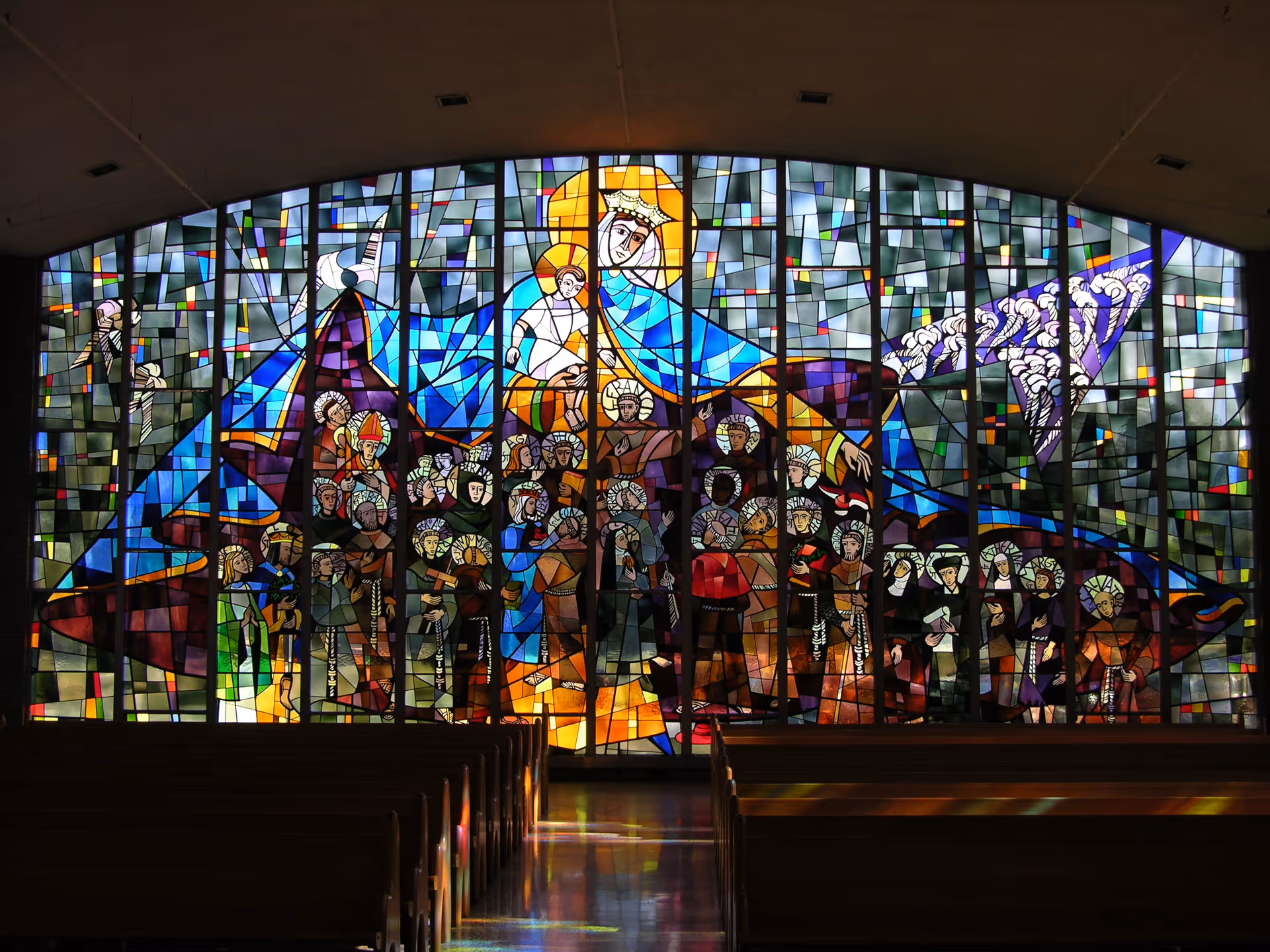A large stained glass window depicting religious figures, including a crowned woman holding a child, surrounded by many other figures with halos, inside a church or chapel with wooden pews in the foreground.