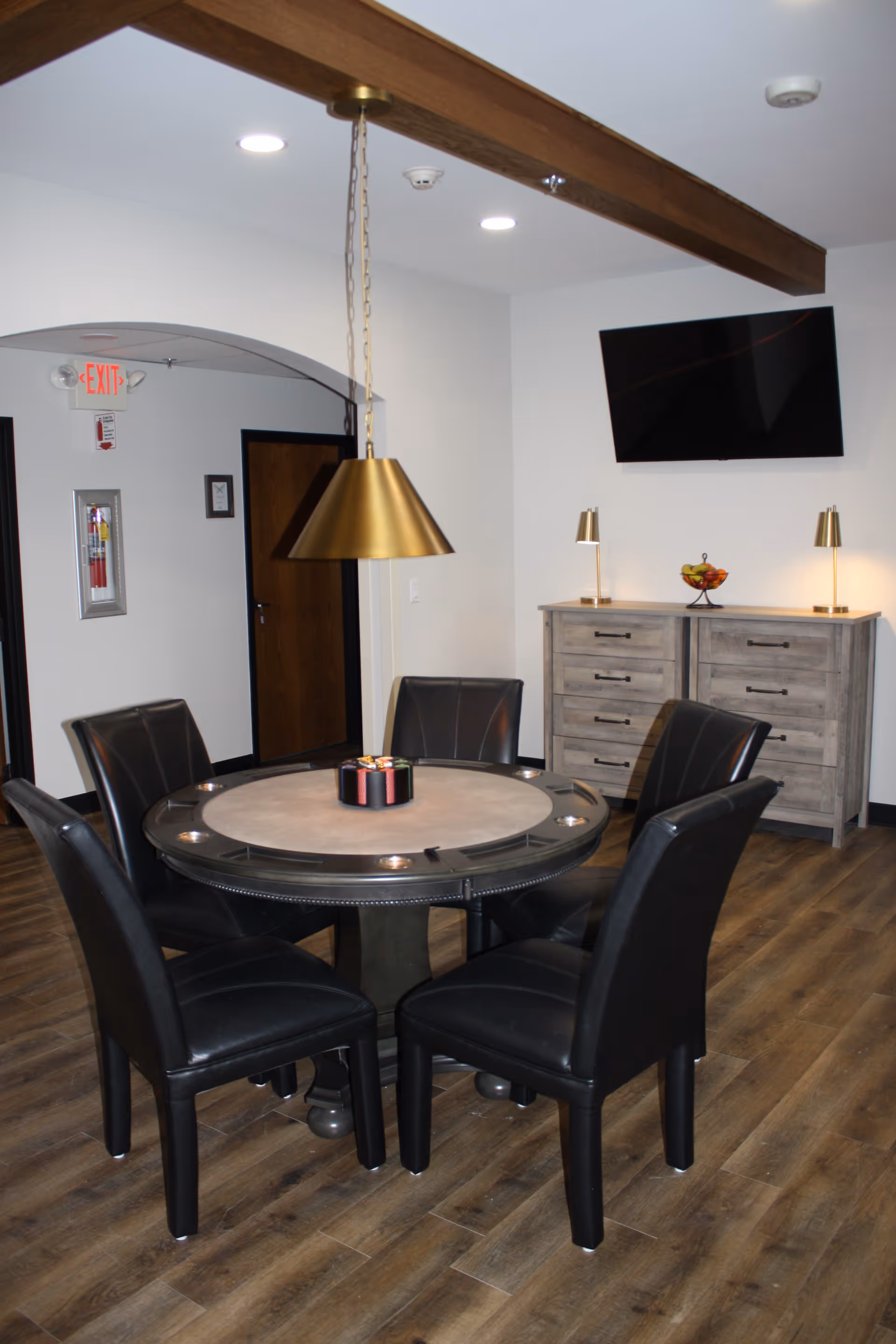 Round poker-style table with six black leather chairs under a brass pendant light, a wall-mounted TV and wooden cabinets in the background.
