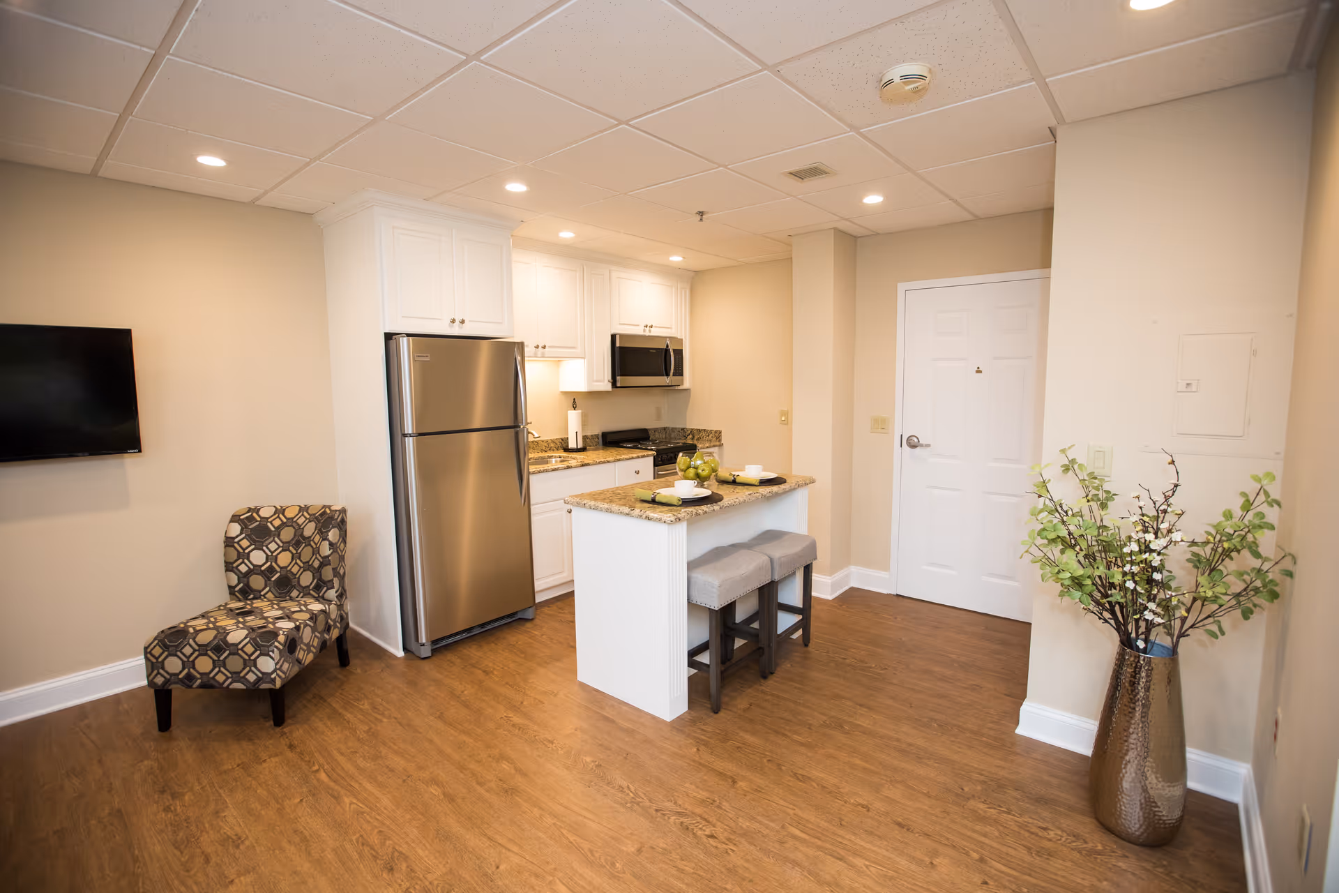 A modern kitchen area in a retirement community featuring white cabinets, a stainless steel refrigerator and microwave, a small island with two stools, a patterned chair, a wall-mounted TV, and a tall vase with green and white flowers on a wooden floor.