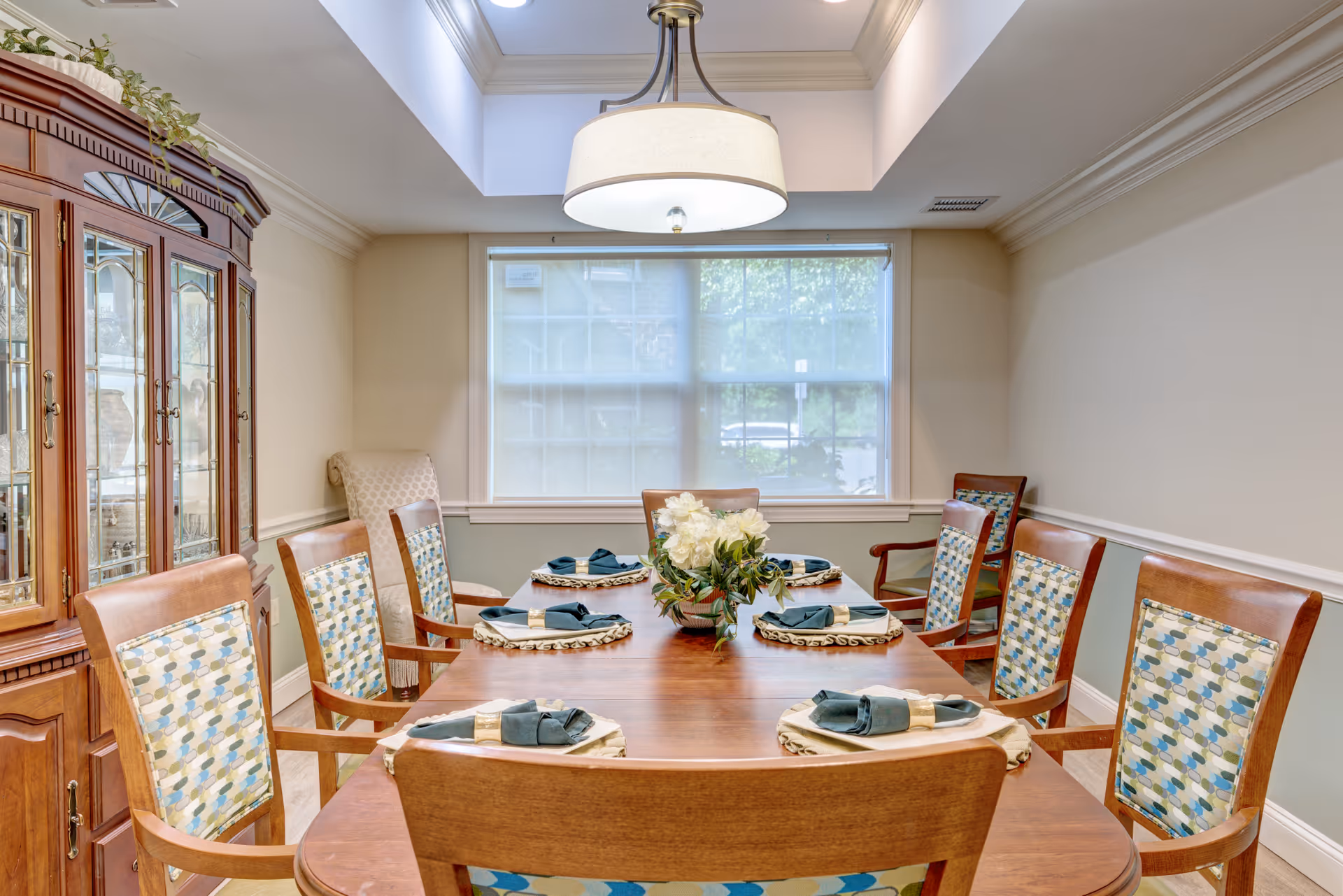 A well-lit dining room with a wooden dining table set for six people. Each place setting has a napkin and a decorative placemat. The chairs have wooden frames with patterned upholstery. A large window with blinds is in the background, and a wooden china cabinet is on the left side of the room. A modern ceiling light fixture hangs above the table.