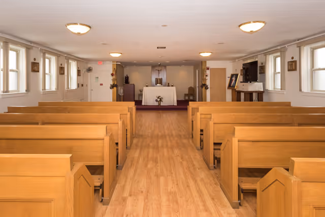 Interior view of a chapel or small worship room with wooden pews arranged in rows facing an altar at the front. The room has light-colored walls, several windows on both sides, ceiling lights, and religious decorations including statues and framed pictures.
