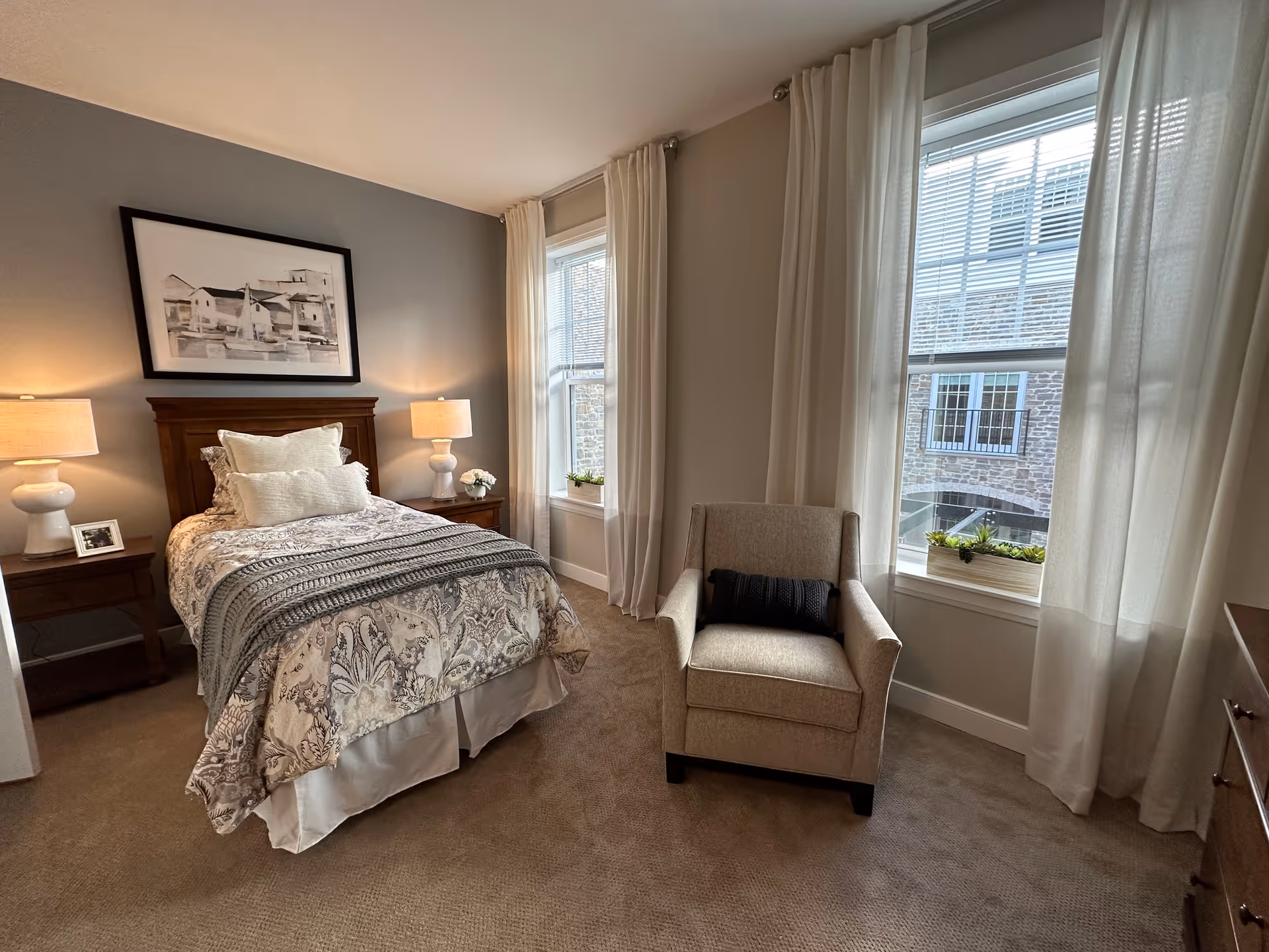 A cozy bedroom with a single bed featuring patterned bedding and multiple pillows. There are two bedside tables with matching lamps on either side of the bed. A framed artwork hangs above the headboard. Next to the bed is a beige armchair with a dark cushion. Two large windows with white curtains allow natural light to fill the room, showing a view of a stone building outside.