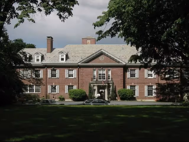 Front exterior view of a large, two-story brick building with white-framed windows, a central entrance with columns, and a gray shingled roof, surrounded by trees and greenery.