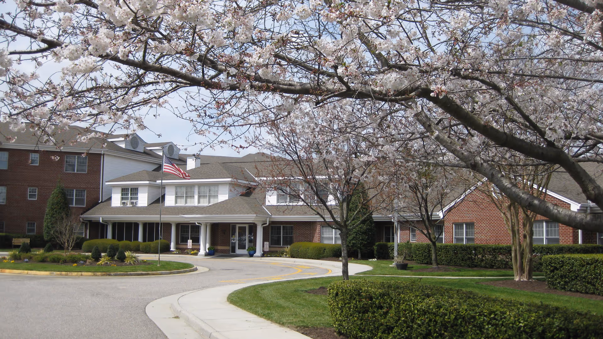 Exterior view of Karolwood Gardens Norfolk Senior Living Center showing a brick building with multiple windows, a covered entrance with white columns, an American flag on a flagpole, and blooming trees in the foreground.