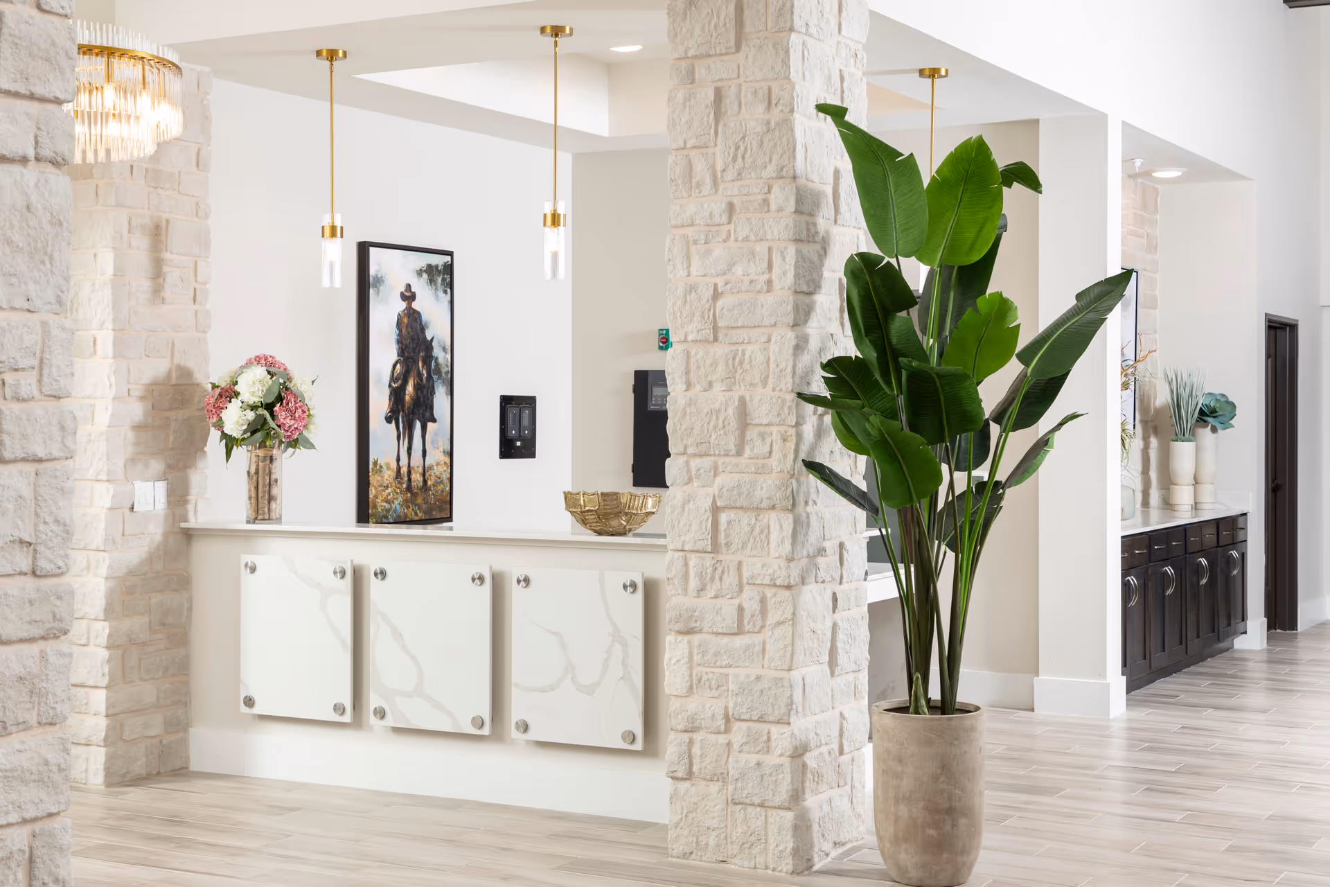 Interior view of a senior living facility featuring a stone pillar, a large potted plant with broad green leaves, a white counter with decorative panels, a vase with flowers, a framed painting of a person on horseback, and modern pendant lights hanging from the ceiling.