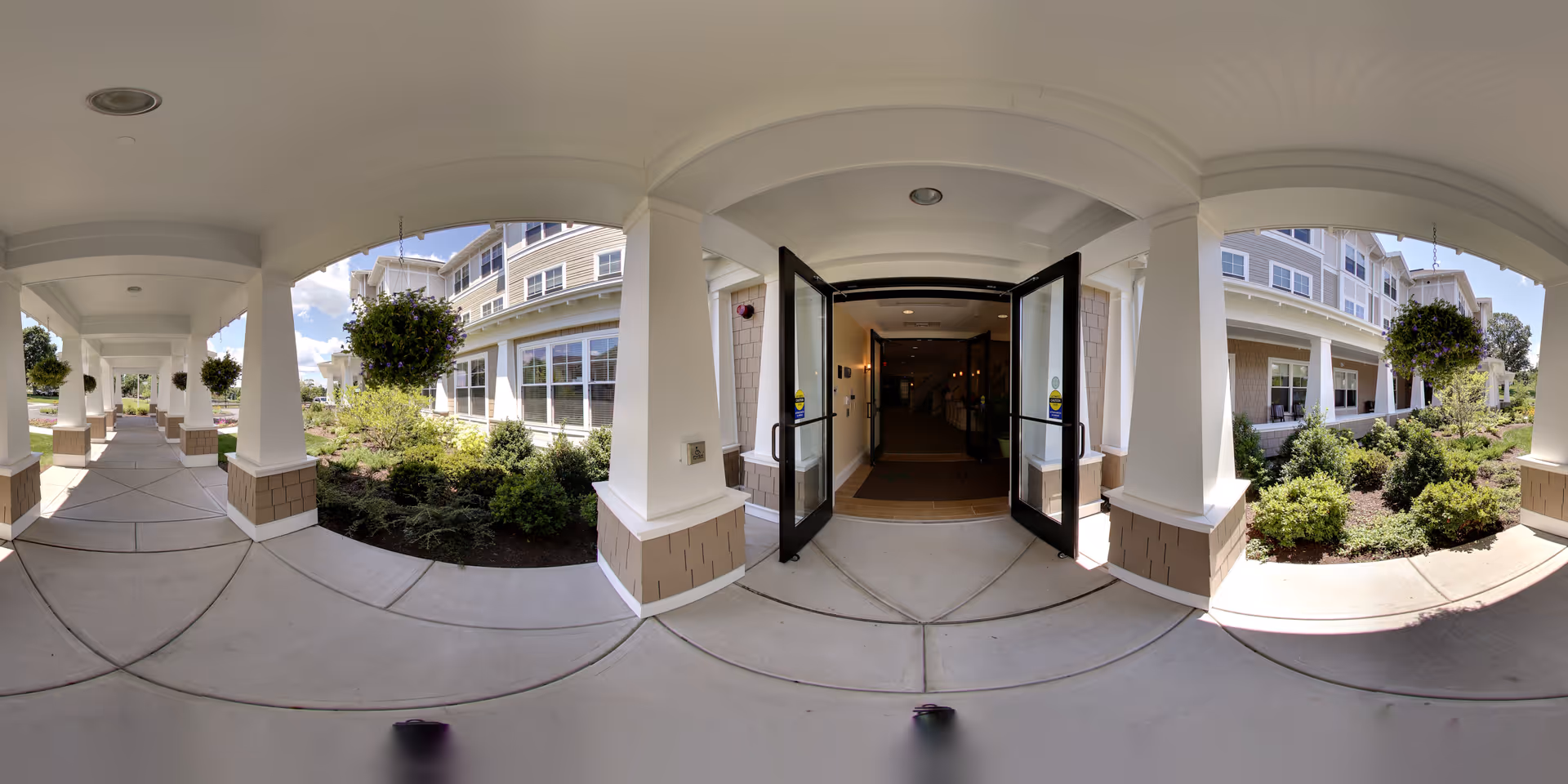 Covered entrance walkway with columns and landscaping leading to open double glass doors of a senior living residence.