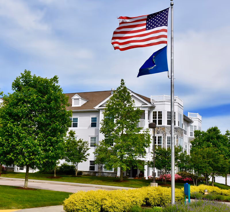 A large white multi-story senior living facility building with stone accents, surrounded by green trees and well-maintained landscaping. Two flags, the American flag and a blue state flag, are flying on a flagpole in front of the building under a partly cloudy sky.