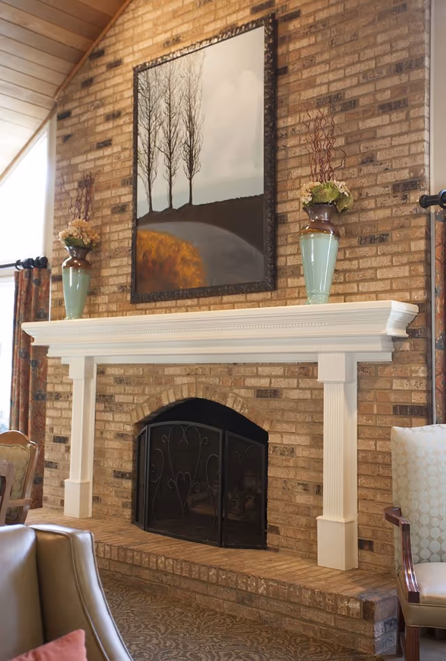 A cozy living room area featuring a brick fireplace with a white wooden mantel. On the mantel are two green vases with floral arrangements. Above the fireplace hangs a painting of four leafless trees on a hill with a cloudy sky. Surrounding the fireplace are upholstered chairs and patterned curtains.