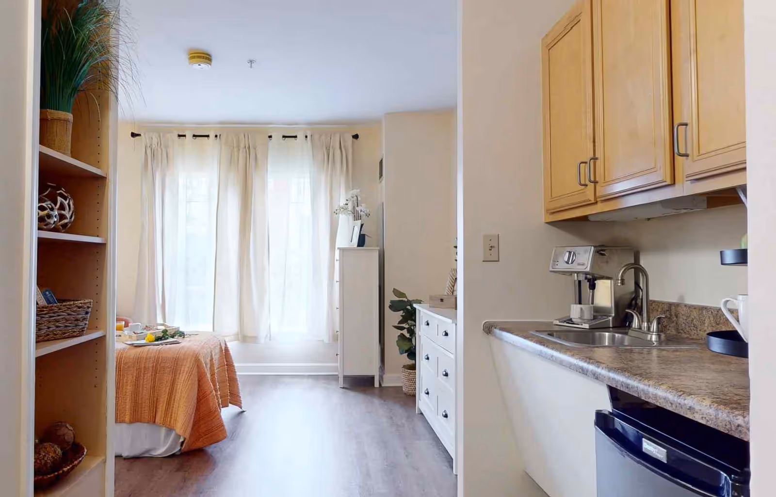A bright and cozy studio apartment with a small kitchenette on the right featuring wooden cabinets, a sink, and a coffee machine. On the left, there is a bed with an orange blanket near a large window with white curtains. A white dresser and some plants are visible in the background, along with a shelving unit on the left side of the image.
