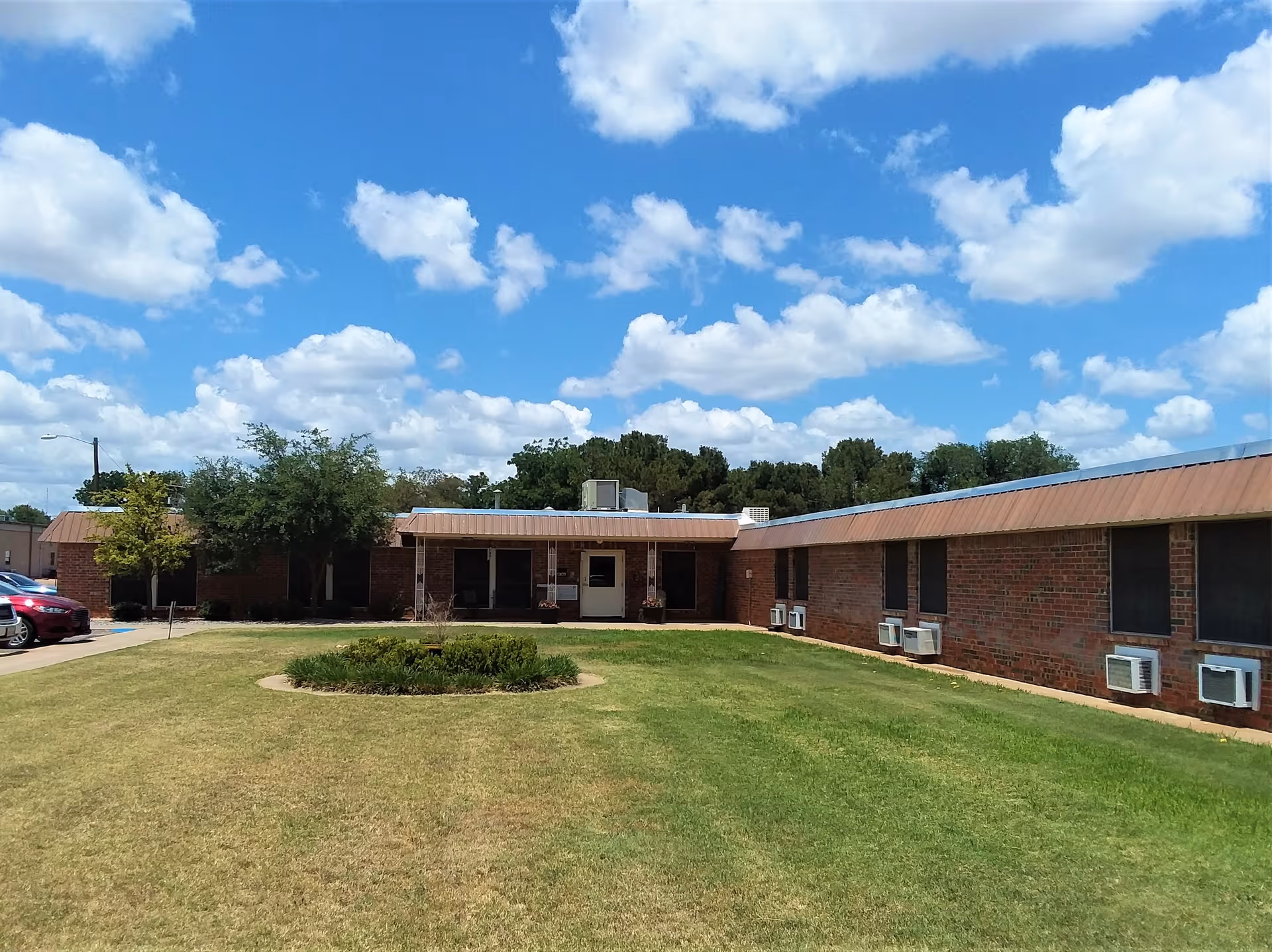 Single-story brick care facility with a grassy front lawn and a circular shrub bed under a blue sky with clouds.