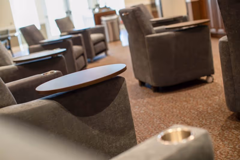 Close-up view of a seating area with several gray upholstered armchairs arranged on a carpeted floor, each chair featuring a small attached wooden table and a built-in cup holder, with natural light coming through windows in the background.