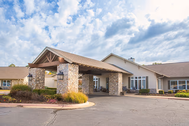 Front entrance of a senior living facility showing a covered porte-cochère with stone pillars, landscaped beds, and the building façade under a partly cloudy sky.