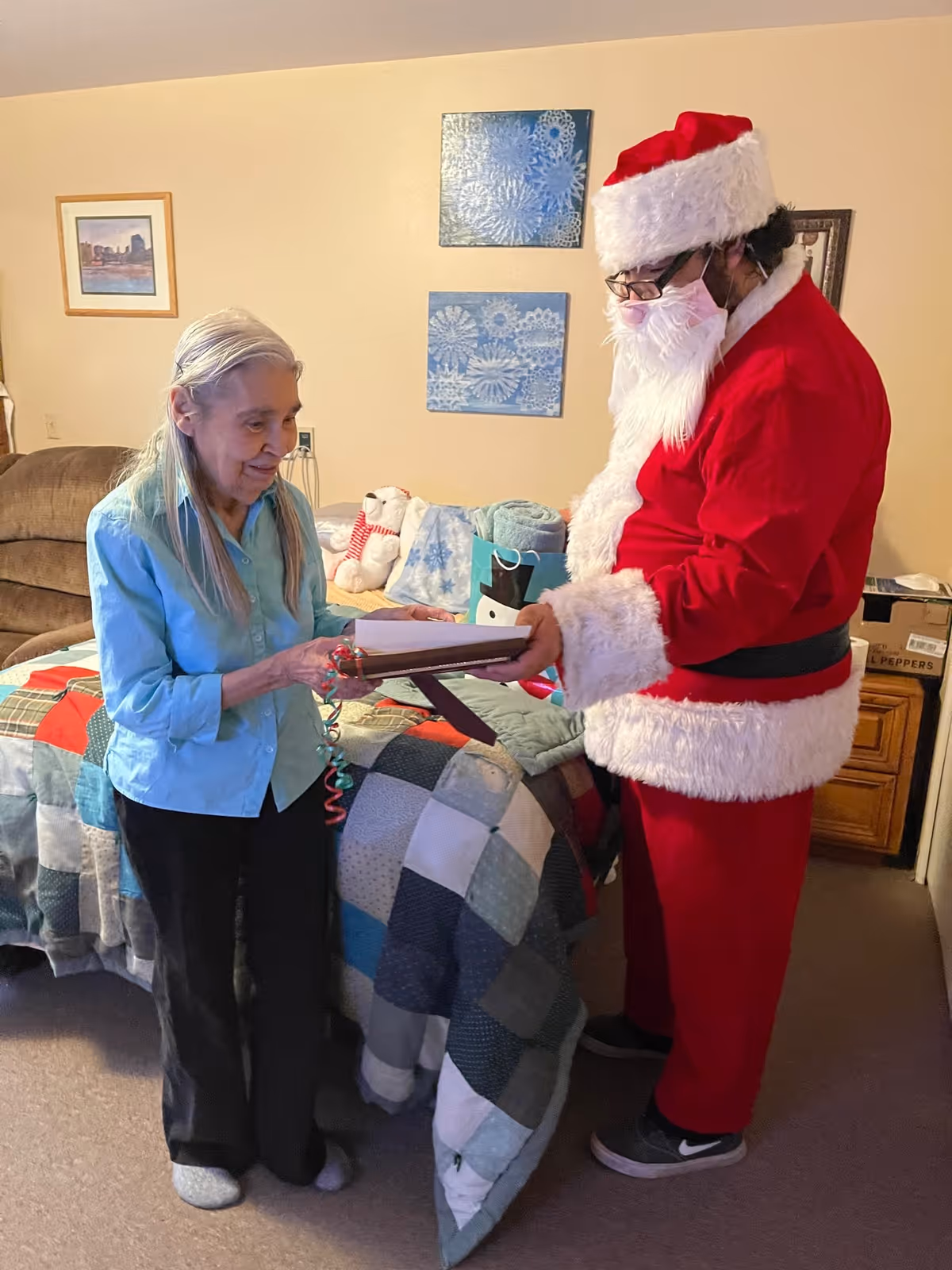 An elderly woman standing next to a bed with a patchwork quilt, receiving a gift from a person dressed in a Santa Claus costume inside a cozy bedroom. The room has framed artwork on the wall and some holiday-themed decorations on the bed.