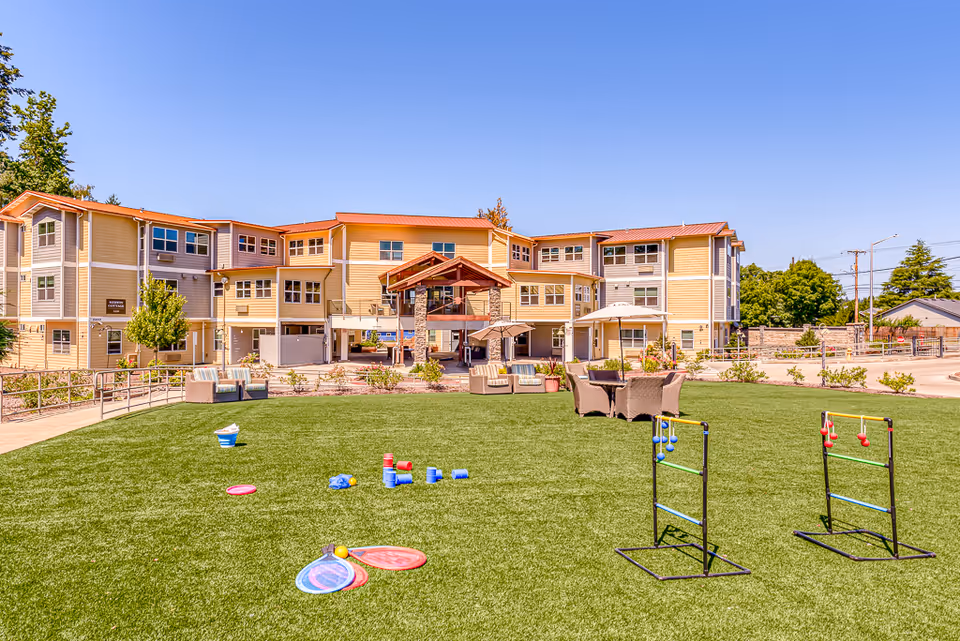 Well-maintained lawn with outdoor games and seating in front of a three-story senior living building under a clear blue sky.