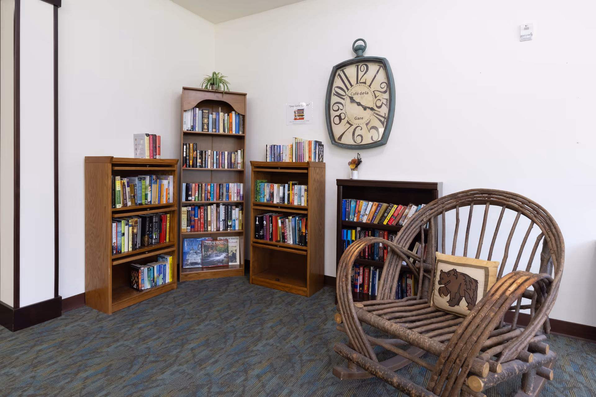 Cozy reading nook with wooden bookshelves filled with books, a rustic wicker chair with a bear pillow, and a large decorative wall clock on the wall.