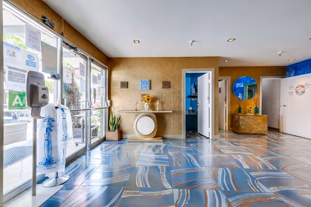 Bright and spacious lobby area with large glass entrance doors on the left, a decorative table with a vase of sunflowers against a gold textured wall, and a blue and gold patterned floor. There are open doorways leading to other rooms and a round mirror on the right wall above a gold cabinet.