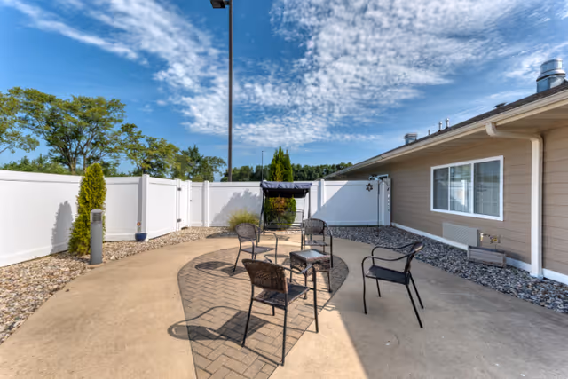 Outdoor patio area with a circular paved section containing four metal chairs and a small table. The patio is enclosed by a white fence and adjacent to a beige building with a window. The sky is partly cloudy with blue patches visible.