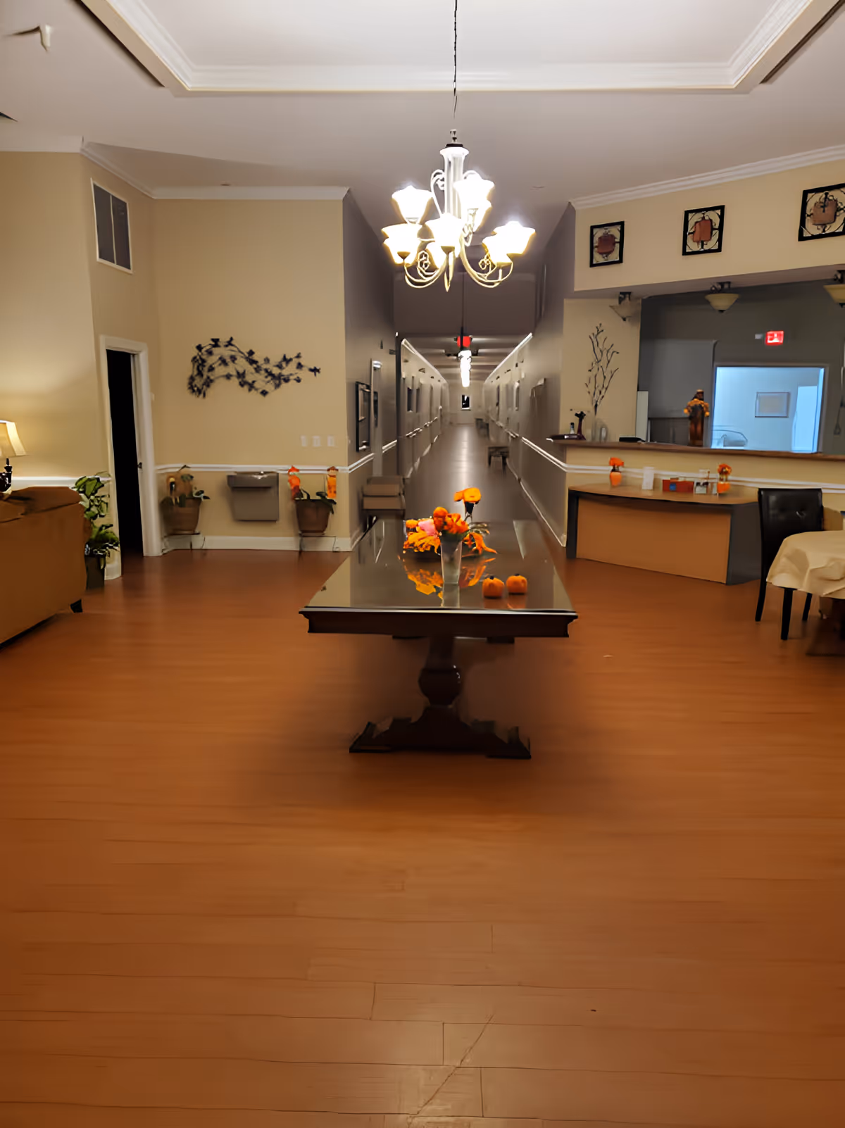 Interior common area of a senior living facility with a long central table decorated with flowers, a chandelier overhead, and a long hallway lined with doors.