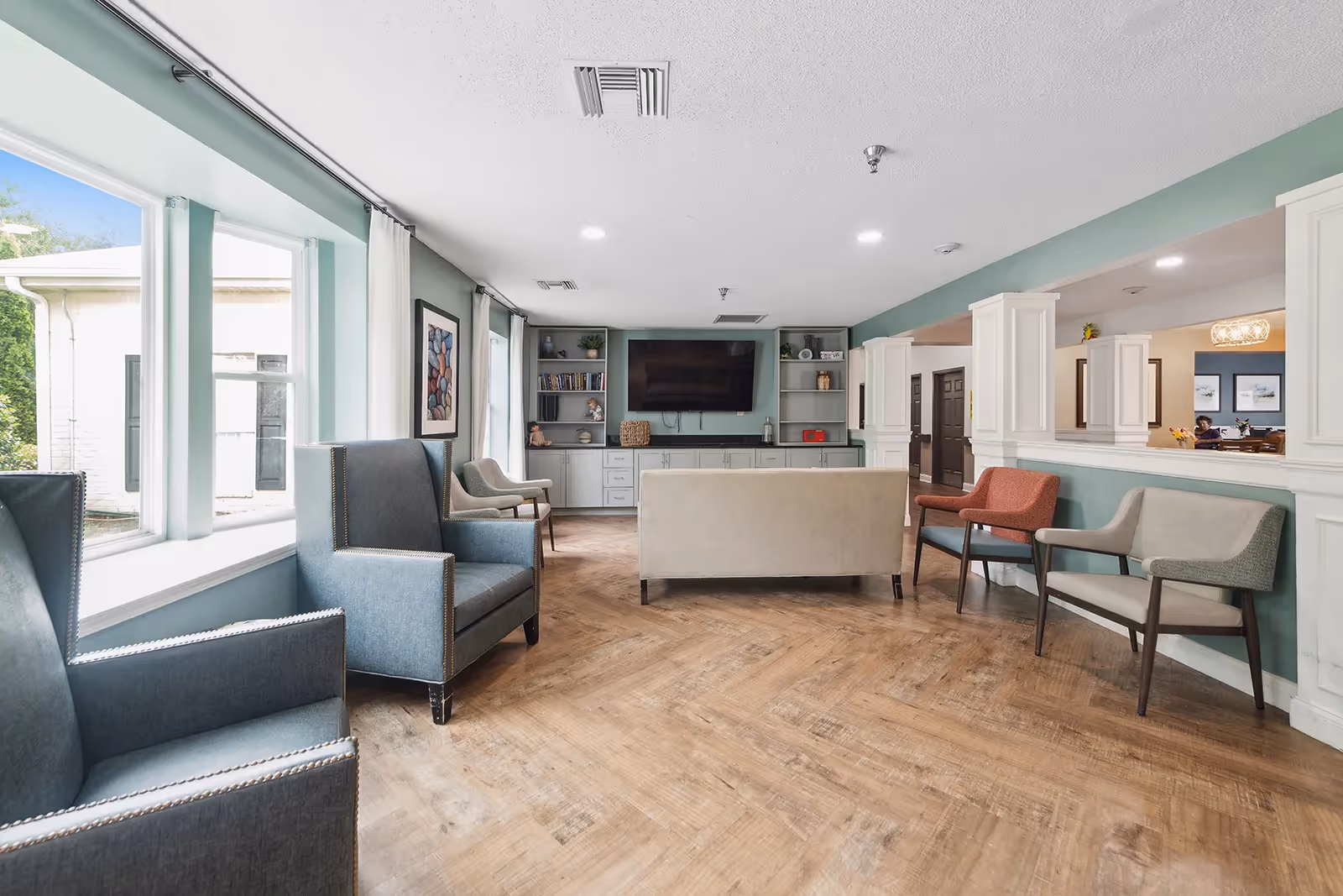 Bright seating area with armchairs, a sofa, built-in shelving and a wall-mounted TV in a memory care community living room.