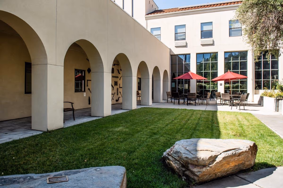 Outdoor courtyard area at The Reutlinger Community featuring a green lawn, large rocks, a building with arched walkways, and patio tables with red umbrellas and chairs.
