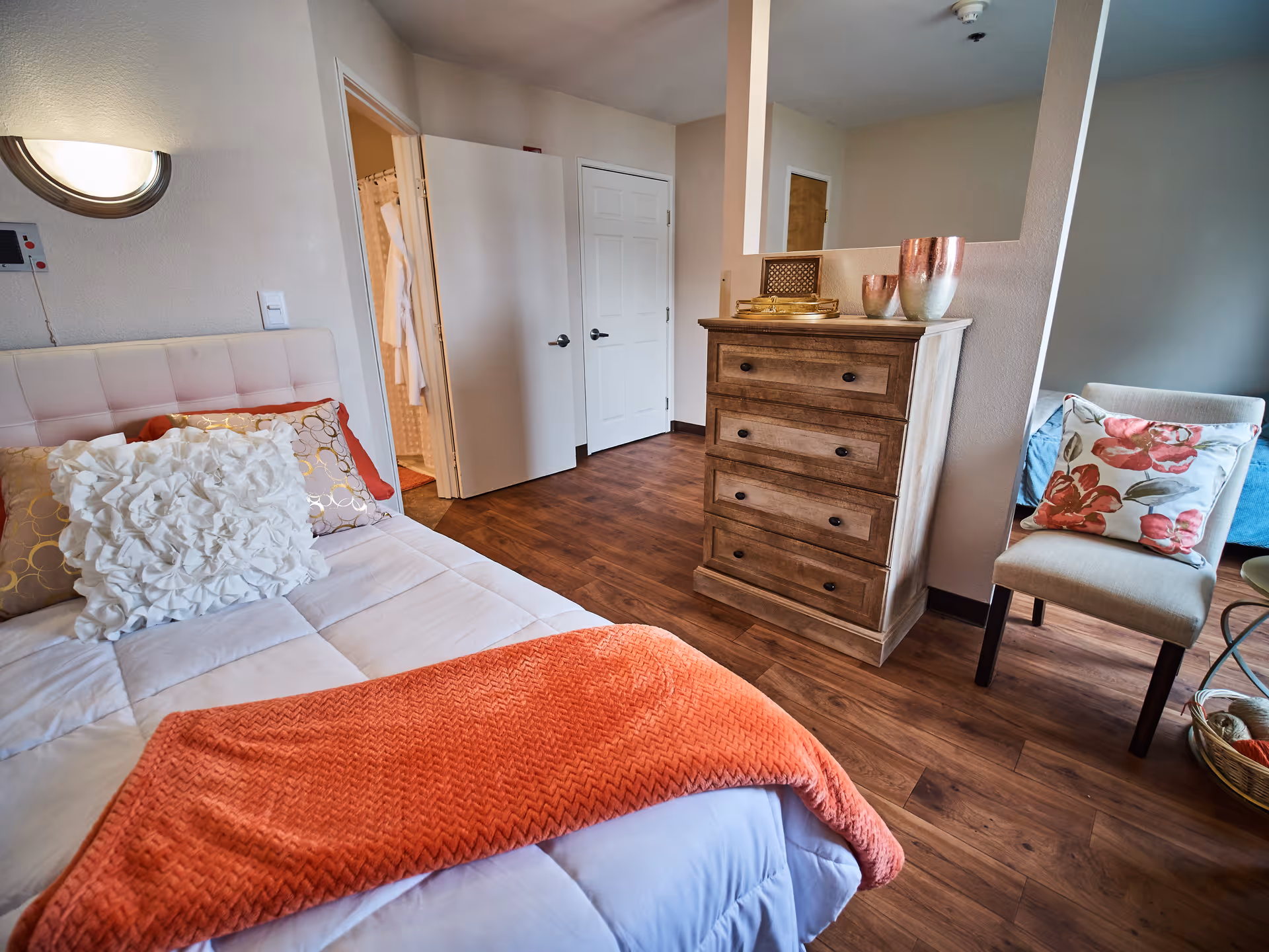 A cozy bedroom in a senior living facility featuring a bed with white and patterned pillows and an orange throw blanket. The room has wooden flooring, a wooden dresser with decorative items on top, a chair with a floral cushion, and an open door leading to a bathroom with a shower curtain visible.
