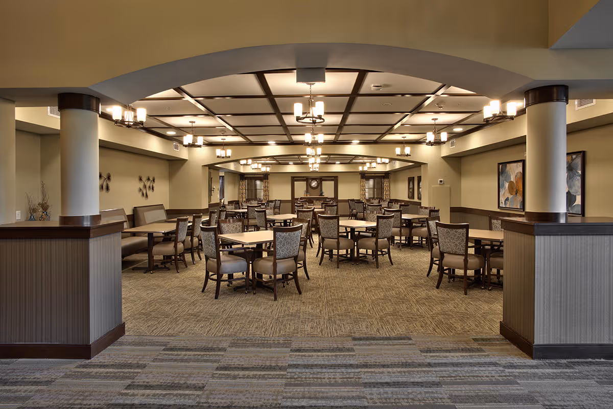 A spacious dining room with multiple square tables and cushioned chairs arranged neatly. The room features a coffered ceiling with several chandeliers providing warm lighting. The walls are decorated with framed artwork and the carpet has a patterned design. Two large columns frame the entrance to the dining area.