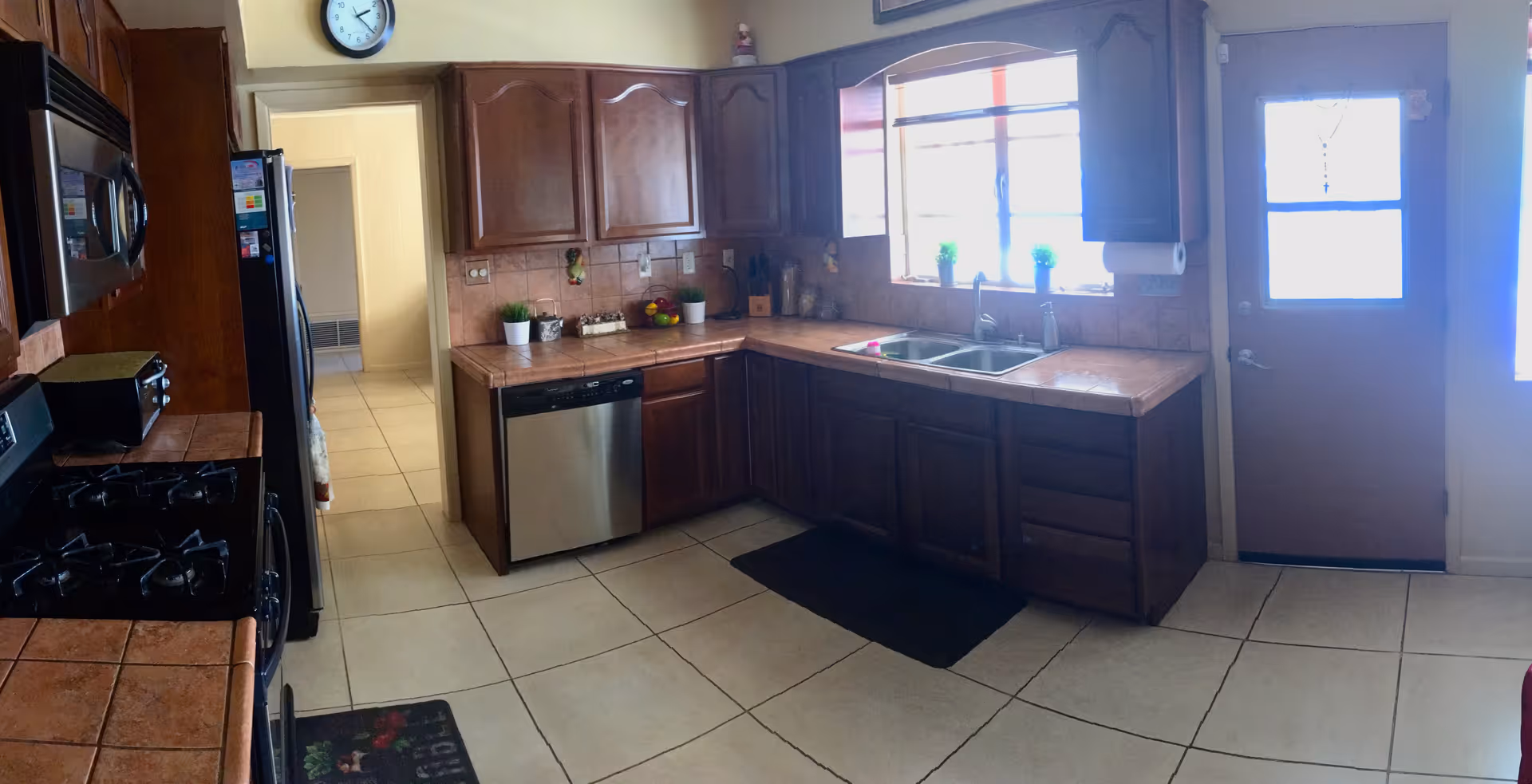 A kitchen with wooden cabinets, tiled countertops, and a tiled floor. The kitchen features a stainless steel dishwasher, a double sink under a window with three small potted plants on the windowsill, a gas stove, a microwave, and a refrigerator. There is a clock on the wall above a doorway, and a door with a window next to the sink area.